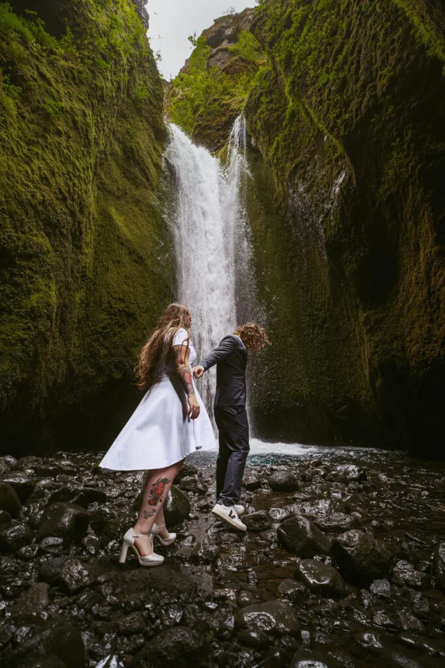 Newlyweds walking barefoot on black rocks beneath the waterfall at Nauthúsagil canyon