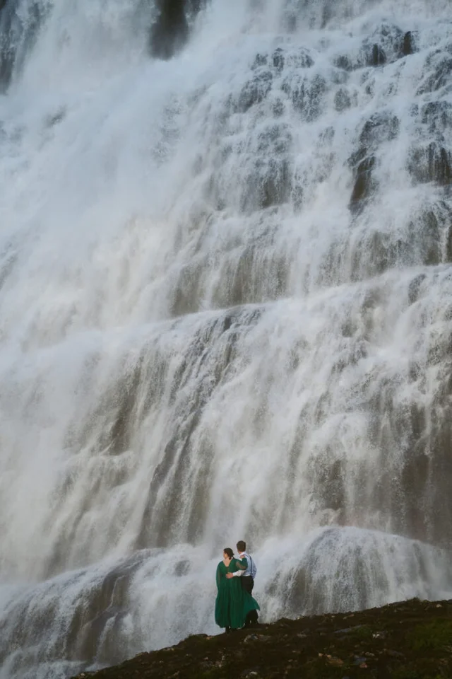 Couple standing at the base of the powerful Dynjandi waterfall, surrounded by mist and rushing water