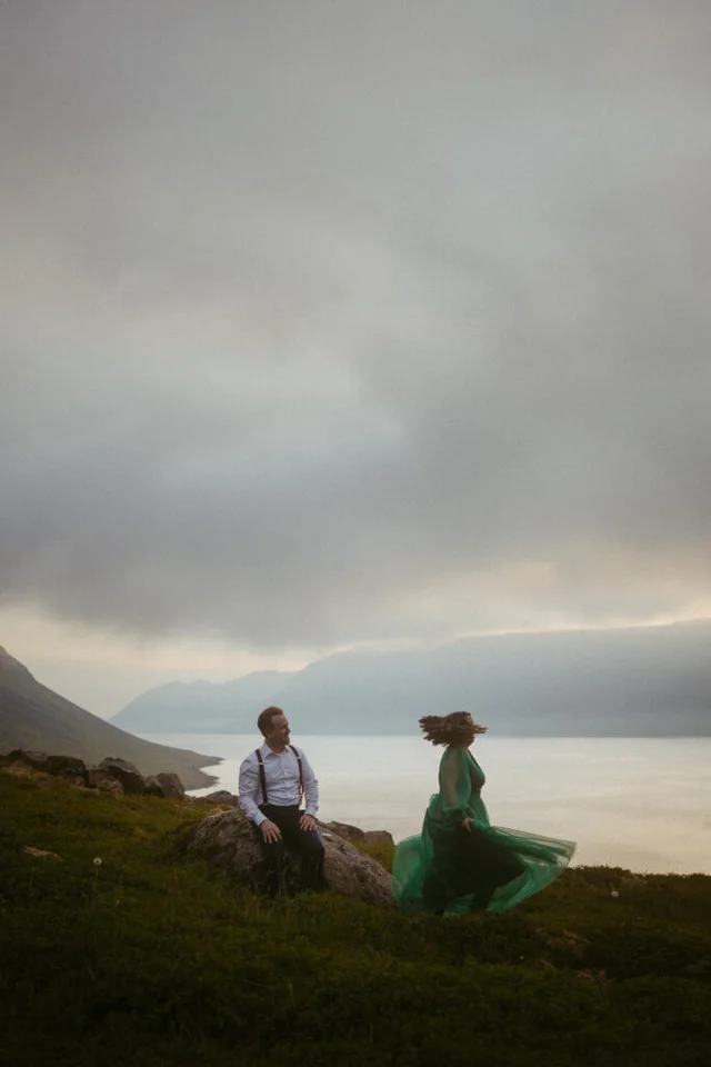 Couple sitting on a grassy hill overlooking a fjord near Dynjandi waterfall in the Westfjords of Iceland