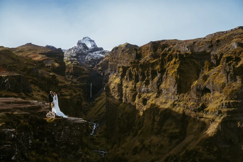 Bride and groom standing on a rocky outcrop above the dramatic cliffs and waterfall of Múlagljúfur Canyon