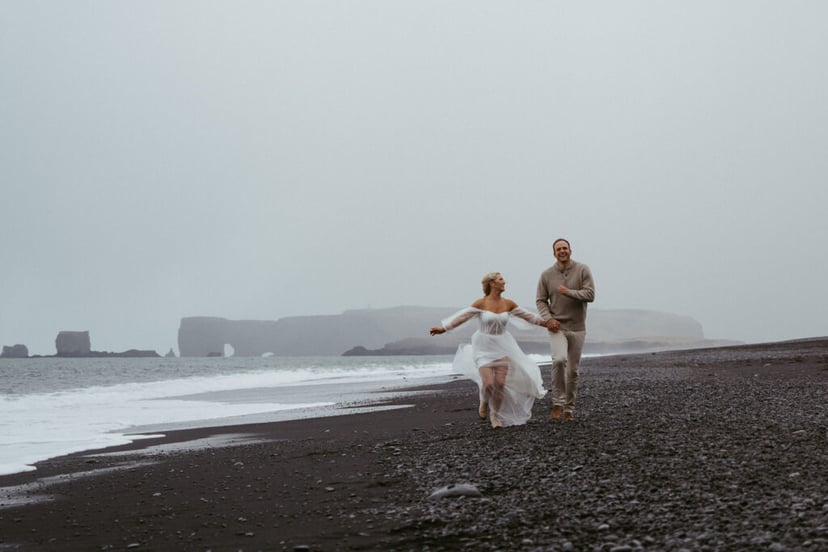 A couple is running in Reynisfjara black sand beach, with Dyrholaey in the background on a moody April day, she has a white dress on celebrating her engagement but could also be a wedding photo