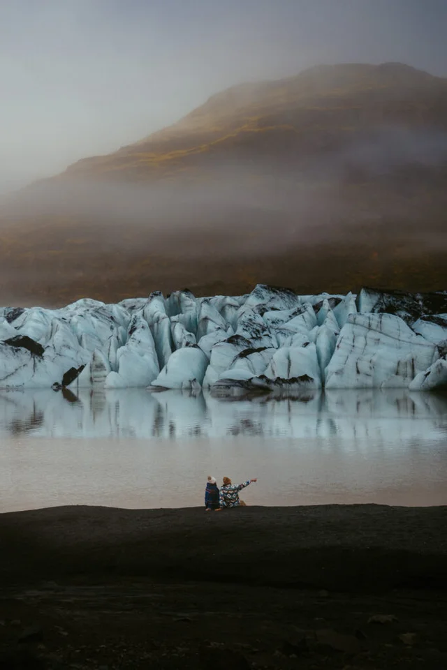 Couple sitting by Sólheimajökull glacier lagoon, pointing at the icebergs under misty morning light