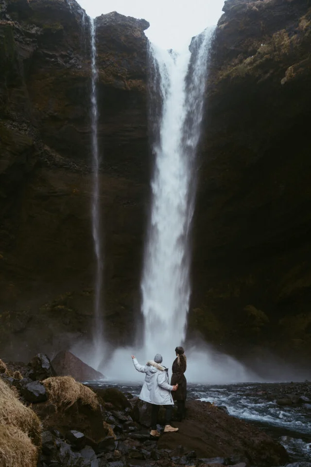 Couple standing near the base of Kvernufoss waterfall in Iceland, looking up at the water