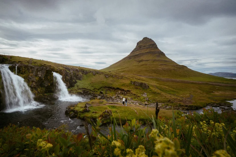 View of Kirkjufellsfoss waterfall with a couple standing on the trail below Kirkjufell mountain in Iceland