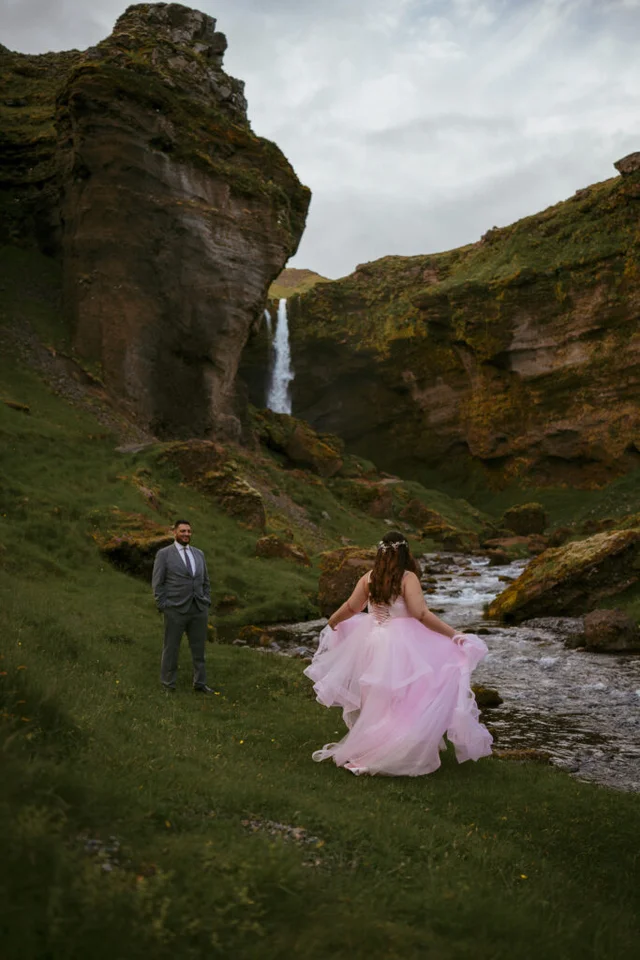 Bride in a pink dress walking across a grassy valley toward Kvernufoss waterfall, with groom waiting ahead