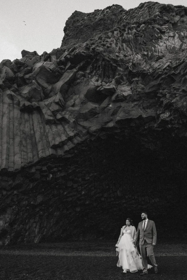 Black and white photo of a couple walking in front of the basalt cave at Reynisfjara beach in Iceland