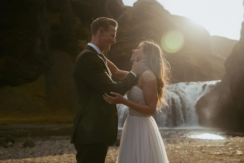 Groom holding bride’s face as they smile in front of Stjórnarfoss waterfall in Iceland during golden hour