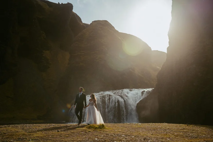 Couple walking hand-in-hand toward Stjórnarfoss waterfall with sunlight glowing behind the cliffs