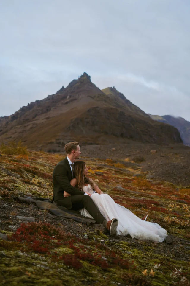 Couple sitting on a mossy hillside near Svinafellsjökull glacier with rugged mountains in the background