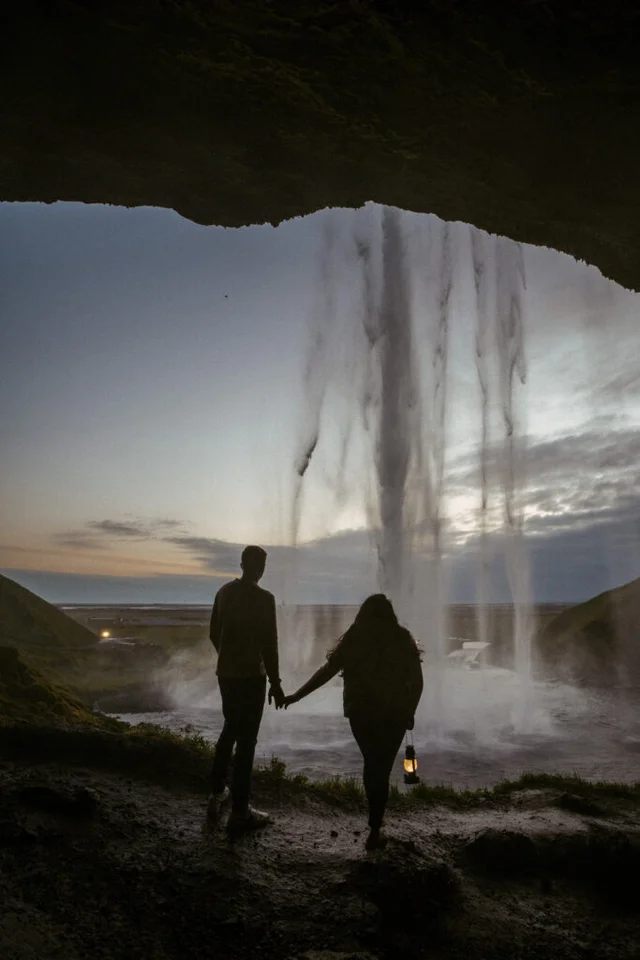 Couple holding hands with a lantern behind Seljalandsfoss waterfall at dusk in Iceland