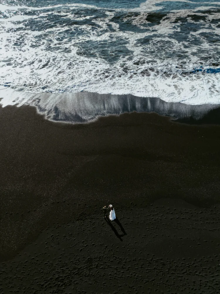 Aerial view of a couple walking along the black sand near the crashing waves at Reynisfjara beach - drone shot