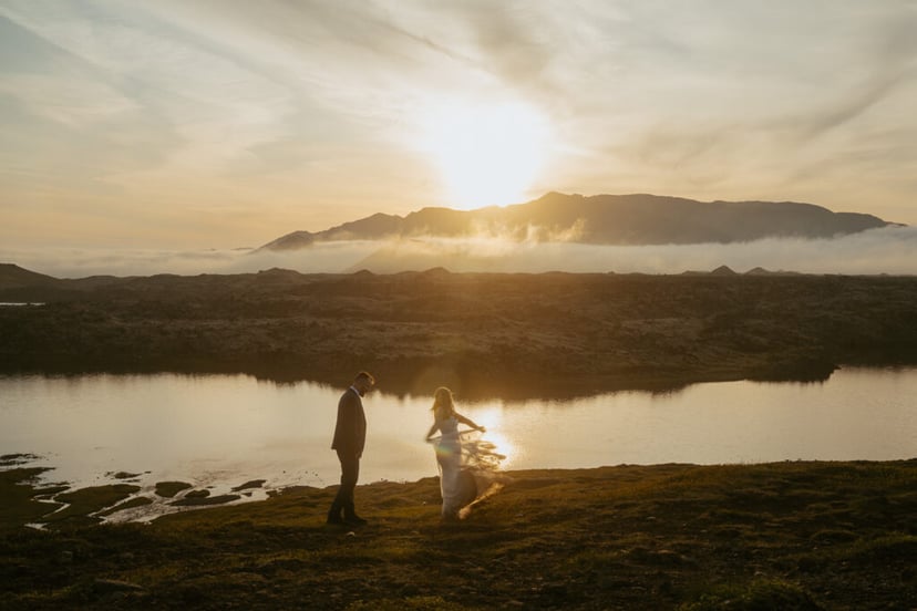 Couple in wedding attire dancing in front of a lake and a view of mountains and fog in the back, sunset