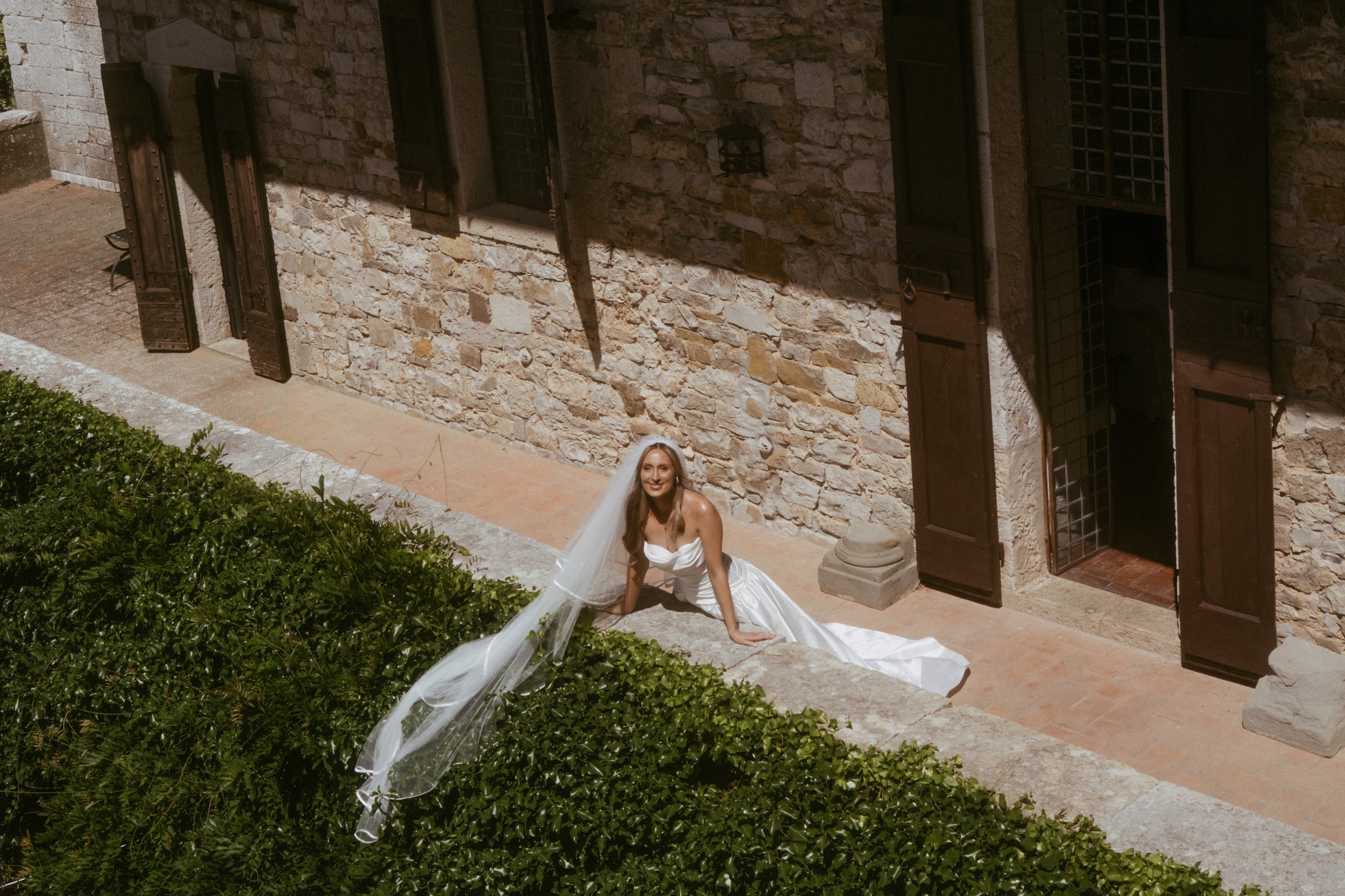 Bride sitting on a stone ledge with her veil trailing over ivy, seen from above, Italy