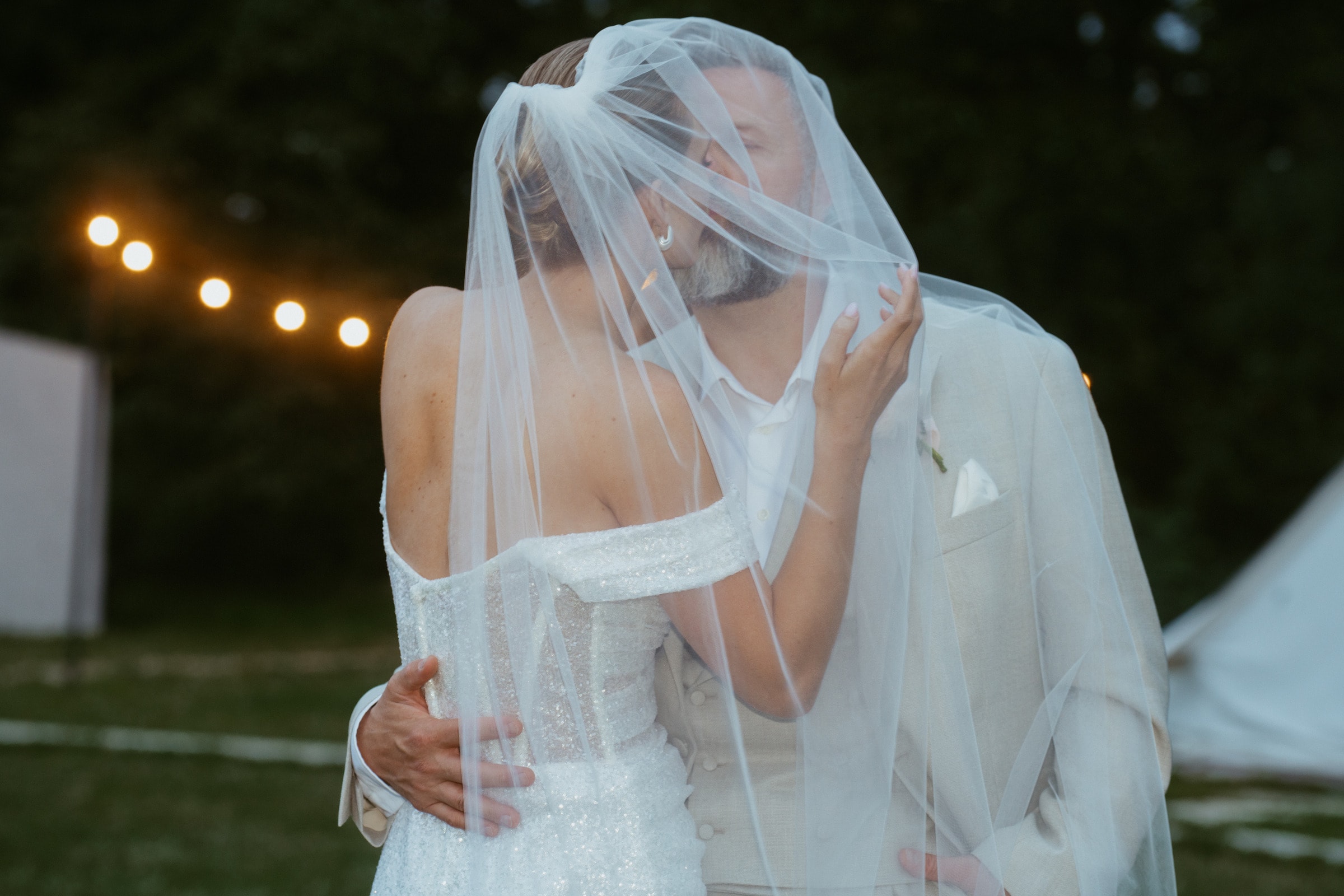Bride and groom kissing under a veil at dusk, string lights in the background, France