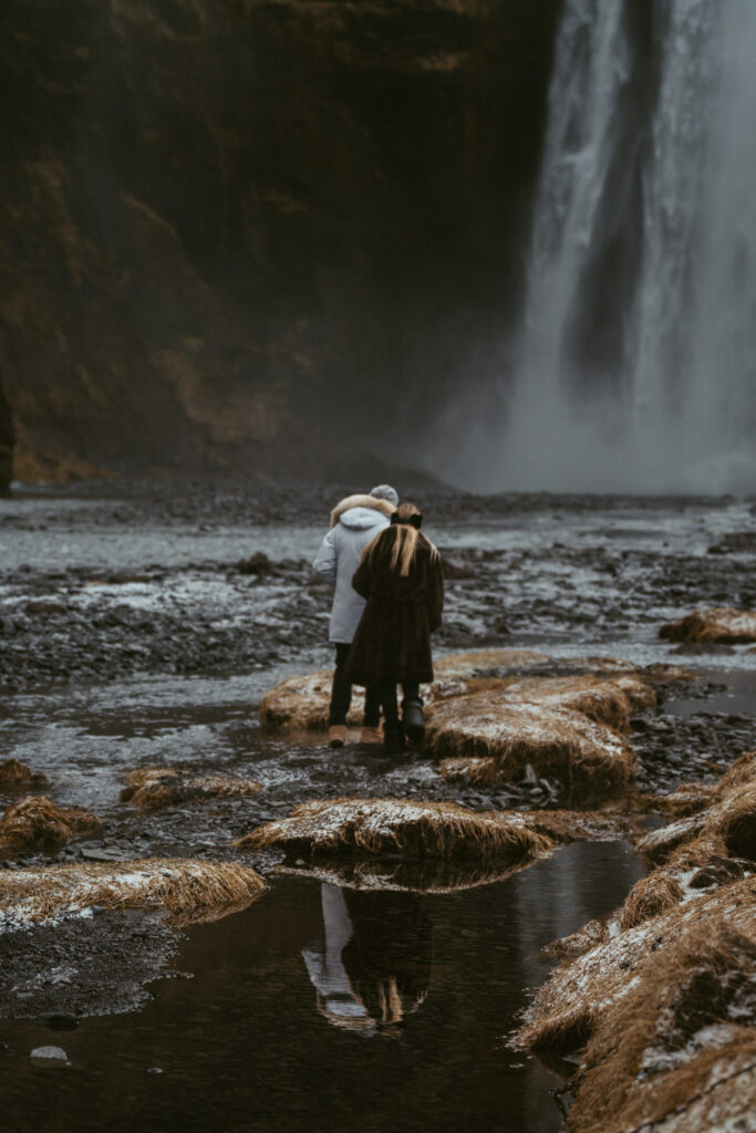 couple embracing in front of Skogafoss waterfall in Iceland after a surprise proposal. The dramatic Icelandic landscape, mist from the falls, and changing weather create a breathtaking backdrop for their engagement session.