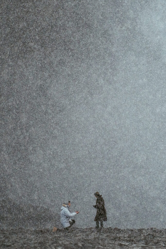 couple embracing in front of Skogafoss waterfall in Iceland after a surprise proposal. The dramatic Icelandic landscape, mist from the falls, and changing weather create a breathtaking backdrop for their engagement session.