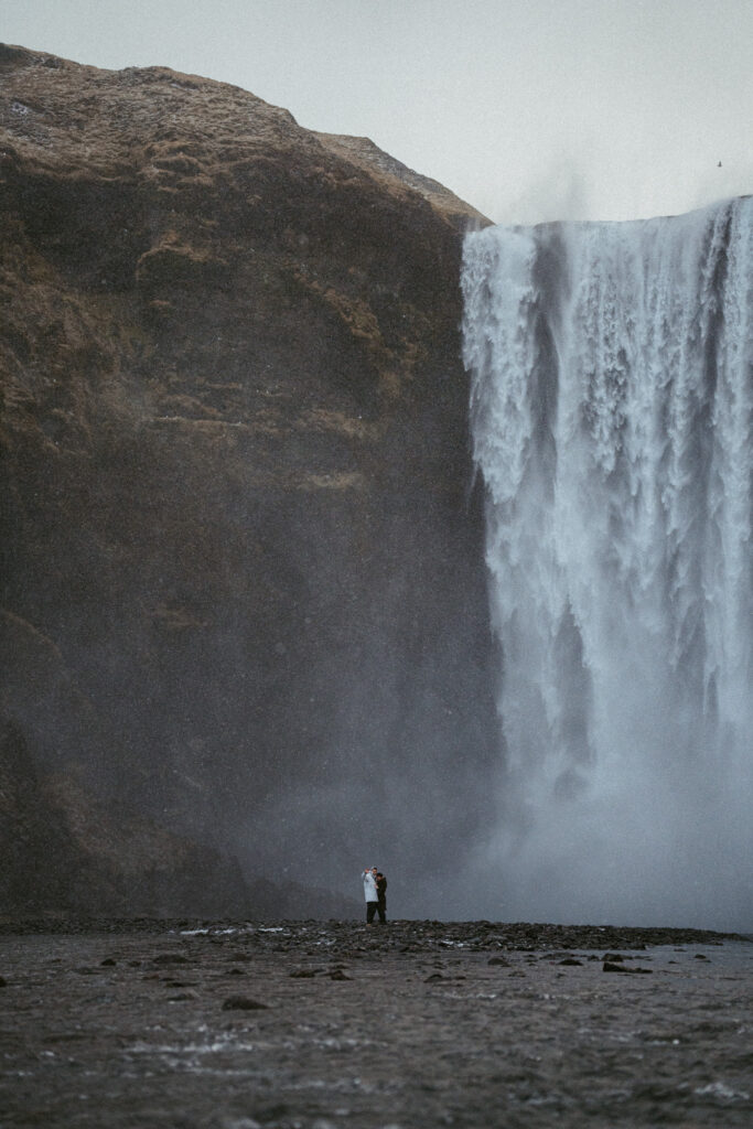 couple embracing in front of Skogafoss waterfall in Iceland after a surprise proposal. The dramatic Icelandic landscape, mist from the falls, and changing weather create a breathtaking backdrop for their engagement session.