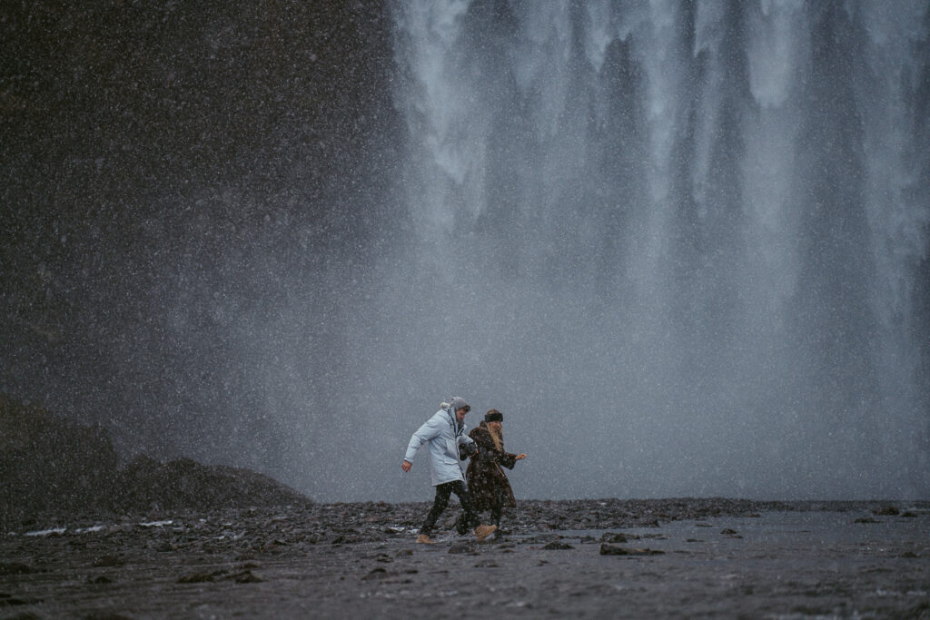 couple embracing in front of Skogafoss waterfall in Iceland after a surprise proposal. The dramatic Icelandic landscape, mist from the falls, and changing weather create a breathtaking backdrop for their engagement session.