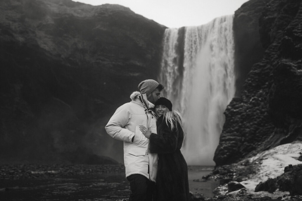 couple embracing in front of Skogafoss waterfall in Iceland after a surprise proposal. The dramatic Icelandic landscape, mist from the falls, and changing weather create a breathtaking backdrop for their engagement session.