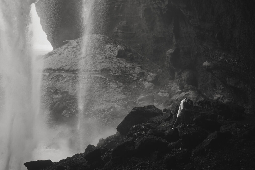 couple embracing in front of Skogafoss waterfall in Iceland after a surprise proposal. The dramatic Icelandic landscape, mist from the falls, and changing weather create a breathtaking backdrop for their engagement session.