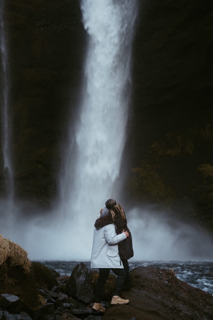 couple embracing in front of Skogafoss waterfall in Iceland after a surprise proposal. The dramatic Icelandic landscape, mist from the falls, and changing weather create a breathtaking backdrop for their engagement session.