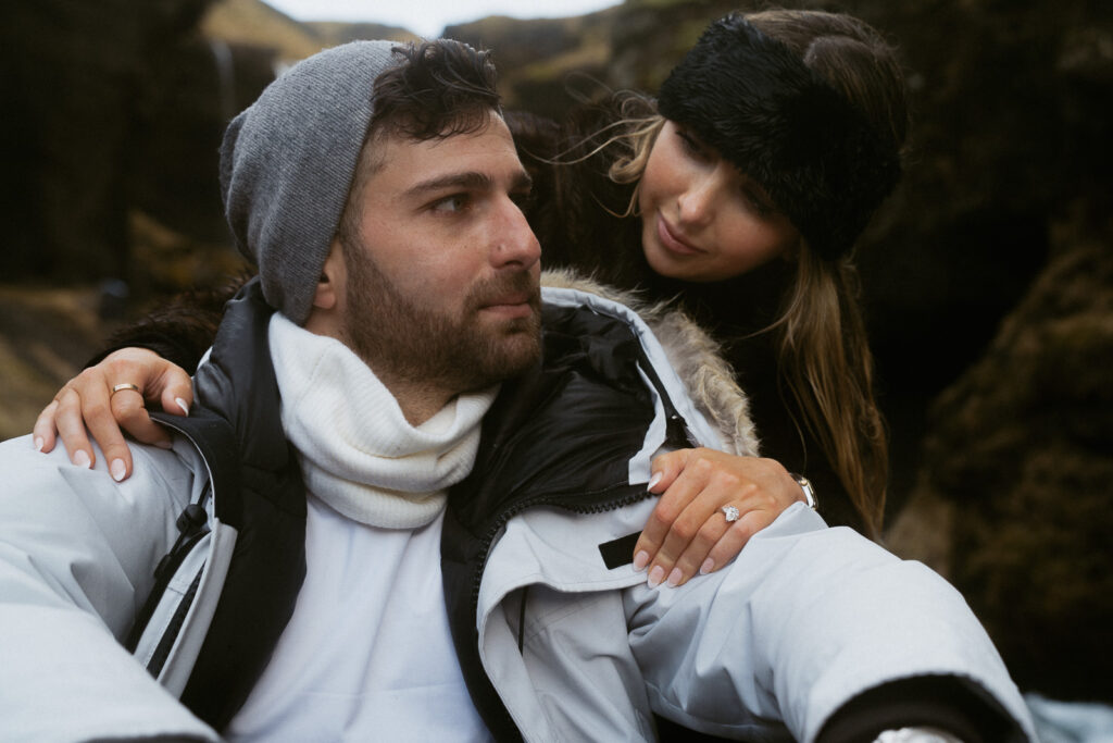 couple embracing in front of Skogafoss waterfall in Iceland after a surprise proposal. The dramatic Icelandic landscape, mist from the falls, and changing weather create a breathtaking backdrop for their engagement session.