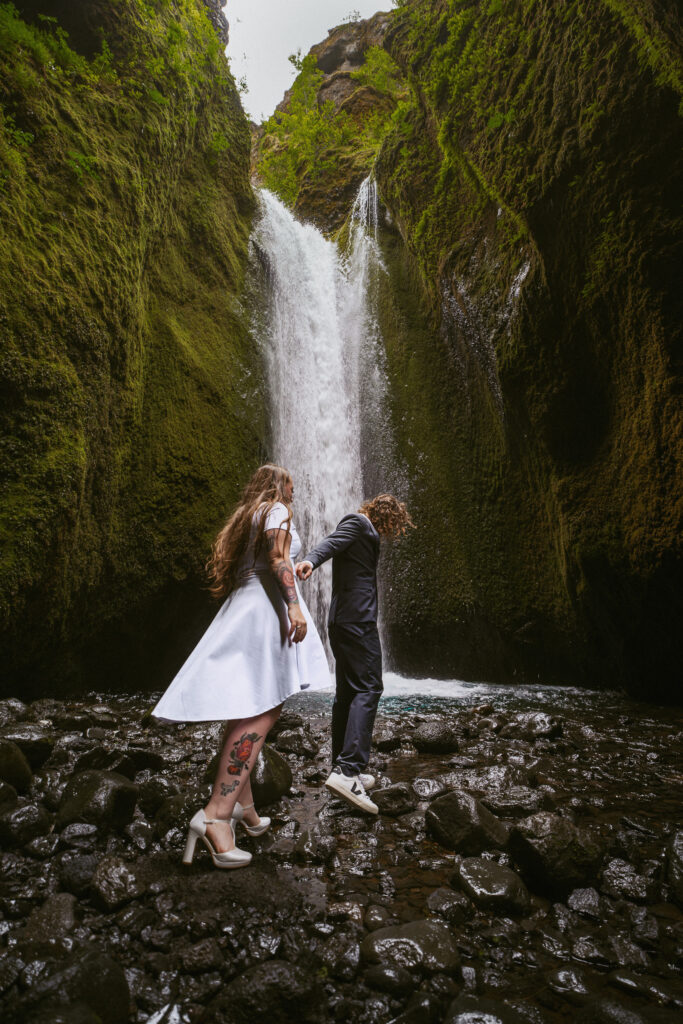 Newlyweds walking barefoot on black rocks beneath the waterfall at Nauthúsagil canyon