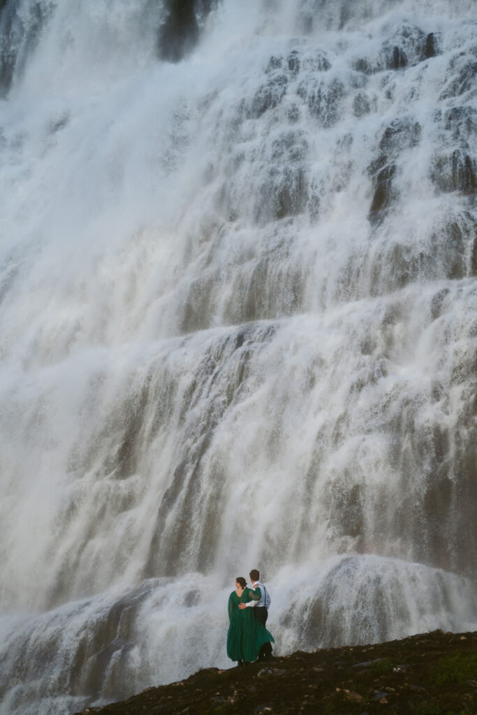 Couple standing at the base of the powerful Dynjandi waterfall, surrounded by mist and rushing water