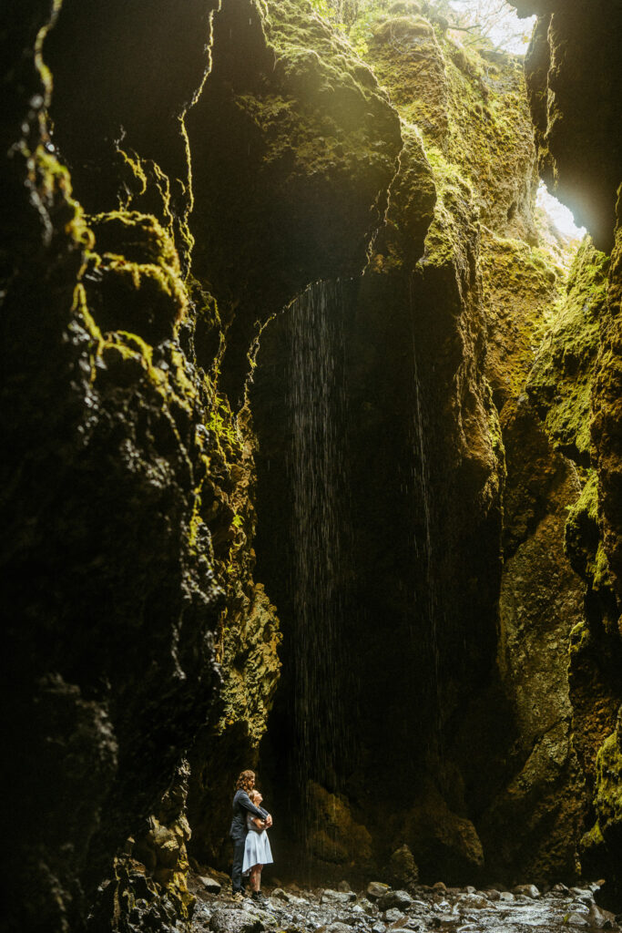Moody light streaming through moss-covered canyon walls with a waterfall and a person standing below at Nauthúsagil