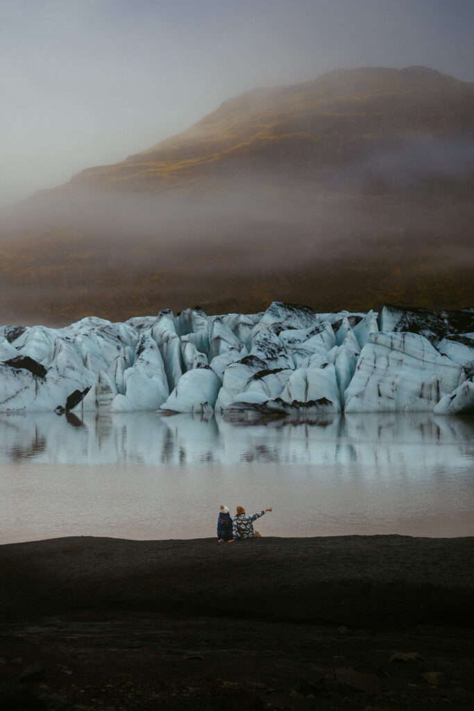Couple sitting by Sólheimajökull glacier lagoon, pointing at the icebergs under misty morning light