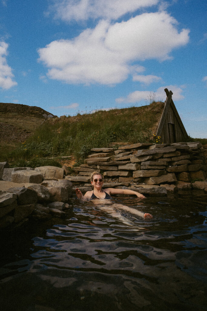 Person relaxing in the stone pool at Hrunalaug hot spring under a blue summer sky in Iceland