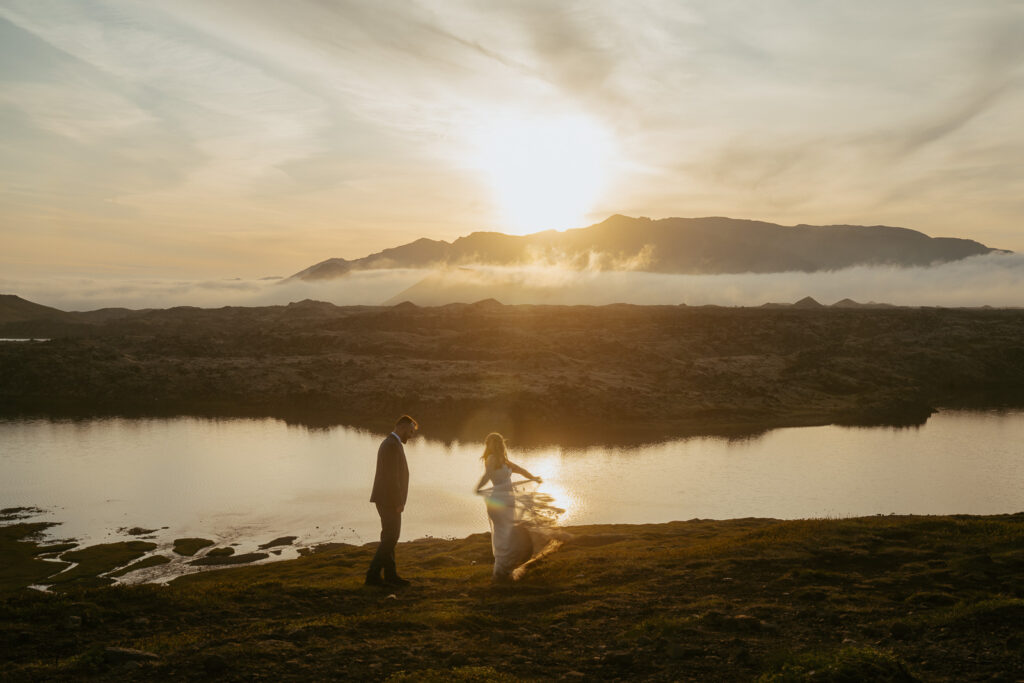 Couple in wedding attire dancing in front of a lake and a view of mountains and fog in the back, sunset