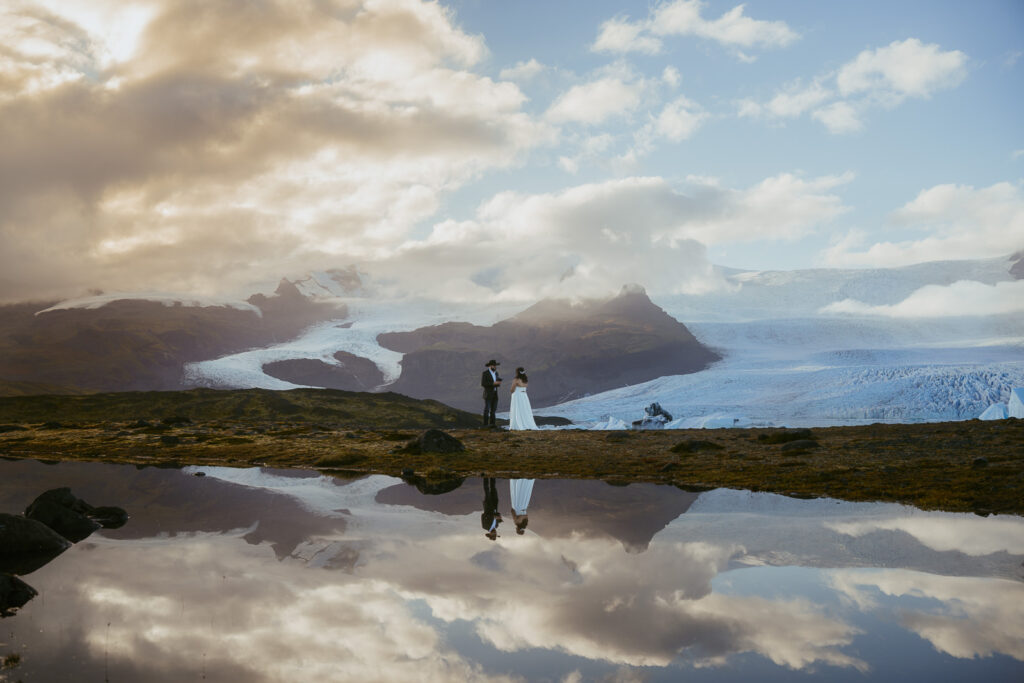 Couple eloping in iceland standing in front of a pond with a reflection. its taking place in front of a glacier.